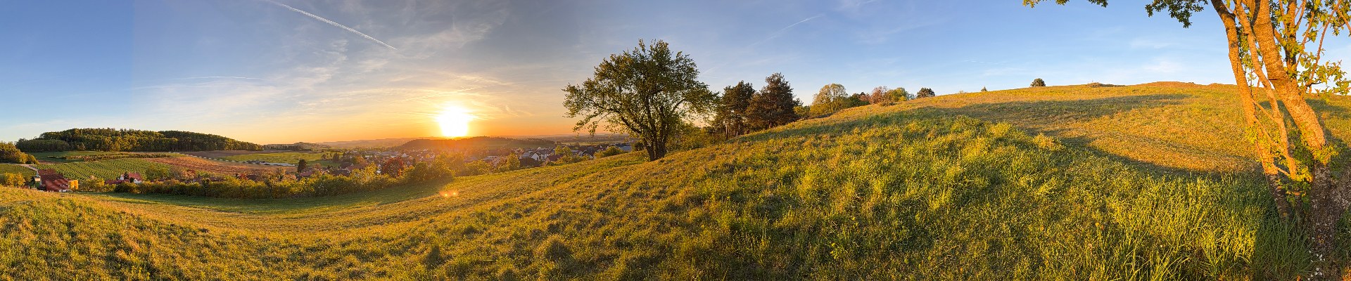 Panorama: Hügelige Wiesenlandschaft mit Ort im Tal bei untergehender Sonne